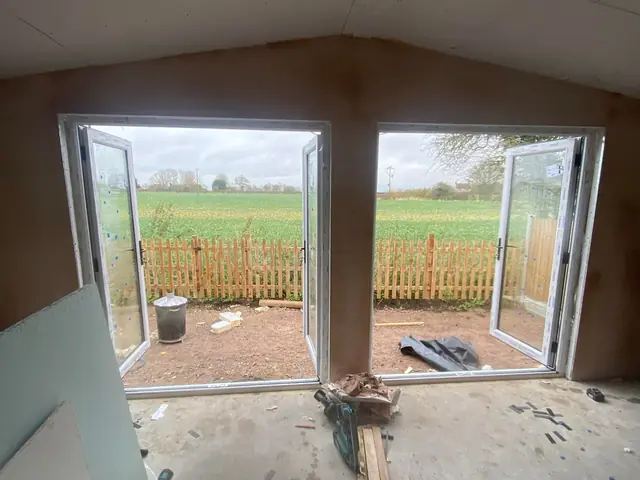 Inside a new extension before plastering, with a concrete floor and open French doors looking out over a fence to a field