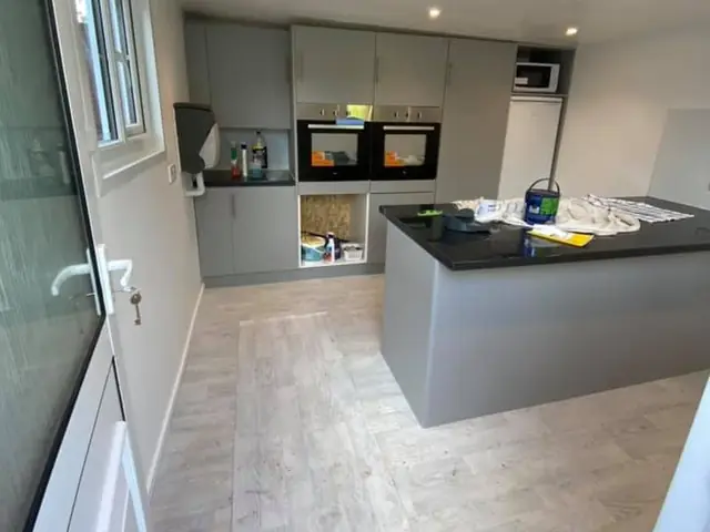 New grey handleless kitchen with two built-in ovens and an island with a black worktop, in a bright room with a light wood-effect floor